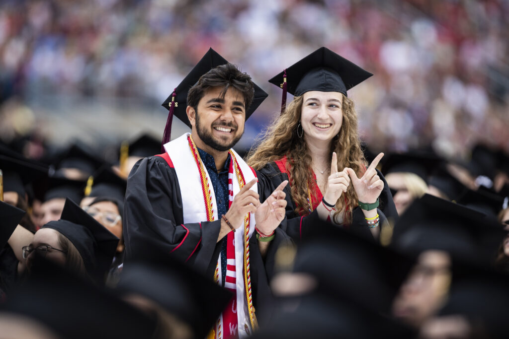 Two graduates making a Wisconsin "W" with their hands, dressed in their cap & gown at commencement